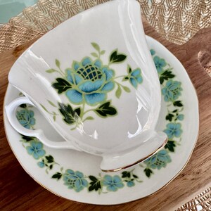 May include: A white teacup and saucer with a floral design. The flowers are blue and green with gold accents. The saucer has a gold rim.