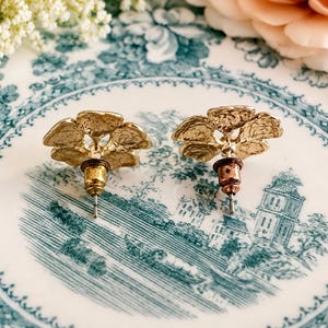 May include: A pair of gold-colored stud earrings, each shaped like a stylized tree or flower. The earrings are set against a vintage blue and white patterned plate, with floral and architectural elements. Soft focus background with flowers.