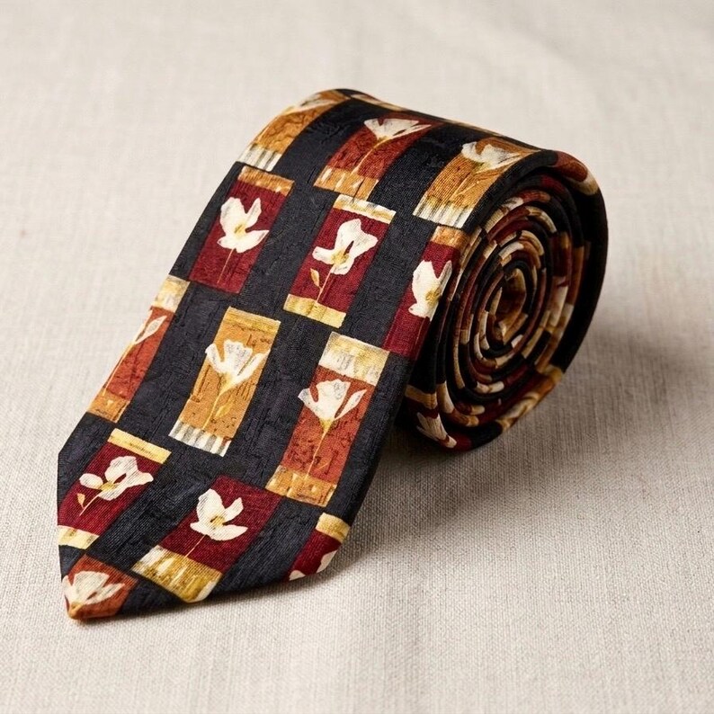 Op de afbeelding: Een stropdas met een patroon met een herhalend bloemmotief. De stropdas heeft een zwarte achtergrond met rechthoekige blokken in tinten rood, goud en bruin. Elk blok bevat een wit bloemmotief. De stropdas is opgerold en toont het patroon.