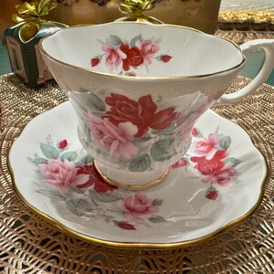 May include: A white teacup and saucer with a floral pattern featuring pink and red roses. The teacup has a gold rim and the saucer has a scalloped edge.