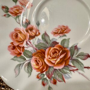 Pode incluir: Um pires de porcelana branca com borda ondulada apresenta um delicado desenho floral de rosas laranja e amarelas com folhas verdes.