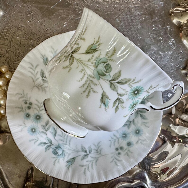 May include: White teacup and saucer set with a floral design in shades of blue and green. The teacup has a curved handle and a gold trim. The saucer has a matching floral pattern. The set is on a silver surface.