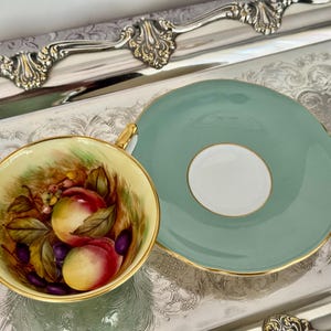 May include: A vintage teacup and saucer set. The teacup features a hand-painted design of peaches and grapes. The saucer is a solid light green color with a gold trim. The set is displayed on a silver tray.