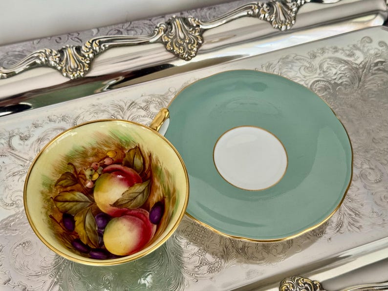 May include: A vintage teacup and saucer set. The teacup features a hand-painted design of peaches and grapes. The saucer is a solid light green color with a gold trim. The set is displayed on a silver tray.