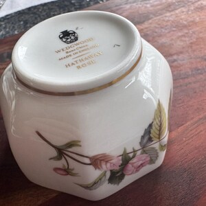 Pode incluir: Uma ch&aacute;vena Wedgwood de cer&acirc;mica branca com um design floral. A ch&aacute;vena tem uma borda dourada e as palavras "WEDGWOOD Bone China MADE IN ENGLAND HATHAWAY ROSE" est&atilde;o impressas na parte inferior.