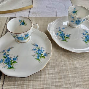 May include: Two white teacups with blue forget-me-not floral designs on a matching set of white plates with gold trim.