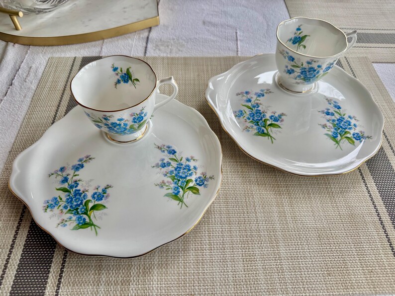 May include: Two white teacups with blue forget-me-not floral designs on a matching set of white plates with gold trim.