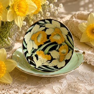 May include: A porcelain teacup and saucer set featuring a black interior decorated with yellow and white daffodil floral patterns. The exterior of the saucer is a pale mint green with a thin gold rim, and the teacup has a gold-trimmed handle.