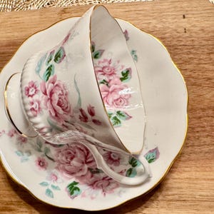 May include: A white teacup and saucer with a floral pattern of pink roses and green leaves. The saucer has a gold rim.