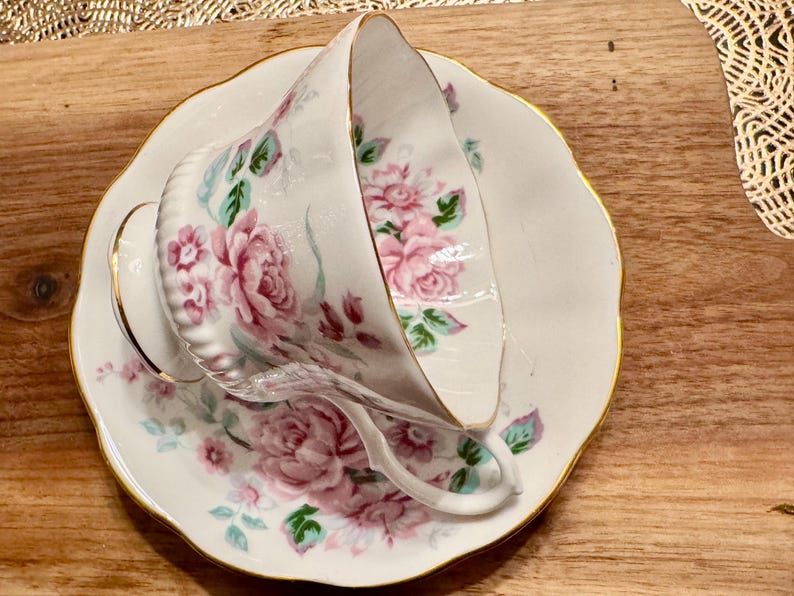 May include: A white teacup and saucer with a floral pattern of pink roses and green leaves. The saucer has a gold rim.