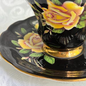 Pode incluir: Um conjunto de ch&aacute;vena e pires de porcelana preta com padr&otilde;es de rosas amarelas vibrantes e folhas verdes. A ch&aacute;vena tem uma borda recortada e uma base dourada, enquanto o pires apresenta uma borda recortada a condizer com um fino acabamento dourado.
