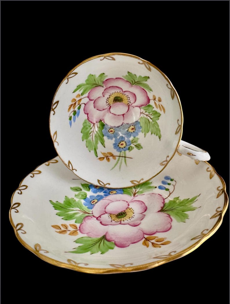 May include: A white teacup and saucer with a pink and blue floral design. The cup and saucer have a gold trim.