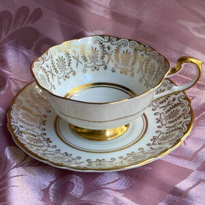 May include: A white porcelain teacup and saucer set featuring a scalloped edge and an ornate gold filigree pattern. The cup has a gold-trimmed handle and a gold base, while the saucer displays a matching intricate gold design along the rim.