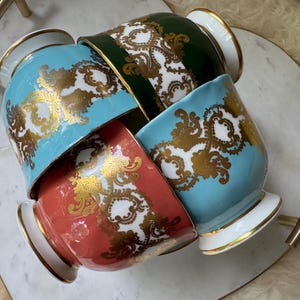 以下が含まれることがあります： ターコイズ、コーラル、グリーンの色合いの装飾用セラミックカップ4つ。それぞれ金と白の華やかなデザインが施されています。カップは、金色のハンドルが付いた白い大理石のトレイに配置されており、豪華な美しさを演出しています。