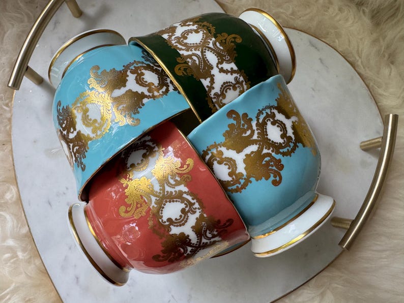 以下が含まれることがあります： ターコイズ、コーラル、グリーンの色合いの装飾用セラミックカップ4つ。それぞれ金と白の華やかなデザインが施されています。カップは、金色のハンドルが付いた白い大理石のトレイに配置されており、豪華な美しさを演出しています。