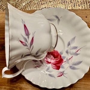 May include: A white teacup and saucer with a delicate floral pattern. The pattern features pink roses and gray leaves on a white background. The teacup and saucer have a gold trim.