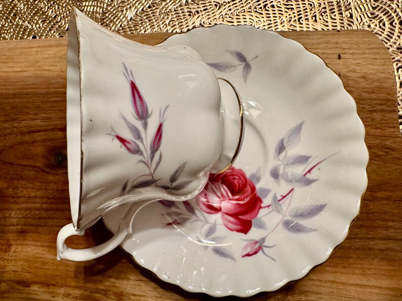 May include: A white teacup and saucer with a delicate floral pattern. The pattern features pink roses and gray leaves on a white background. The teacup and saucer have a gold trim.