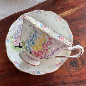 May include: A white teacup and saucer with a floral design. The cup and saucer are decorated with colorful swirls and dots. The cup has a gold rim.