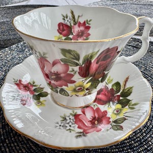 Puede incluir: Un juego de taza y platillo de t&eacute; blanco con un patr&oacute;n floral con rosas rosas, rojas y amarillas. El juego tiene un borde dorado.