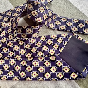 Puede incluir: Una corbata de color morado oscuro con un patr&oacute;n geom&eacute;trico repetitivo de l&iacute;neas blancas punteadas. Cada cuadrado de la cuadr&iacute;cula contiene un motivo floral estilizado de color crema con un peque&ntilde;o centro rojo. La corbata tiene un forro negro liso.