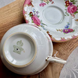 May include: A white teacup and saucer set with a floral design featuring pink roses and other colorful flowers. The teacup has a gold handle and trim. The saucer has a pink and gold border. The set is on a wooden surface.