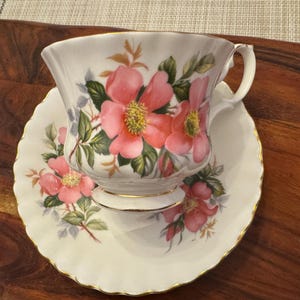 Puede incluir: Un juego de taza y platillo de t&eacute; blanco con un dise&ntilde;o floral con rosas rosas y hojas verdes. La taza de t&eacute; tiene un borde dorado y el platillo tiene un borde festoneado.