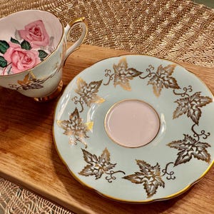 May include: A light blue teacup and saucer set with a gold leaf design. The teacup features a pink rose pattern.