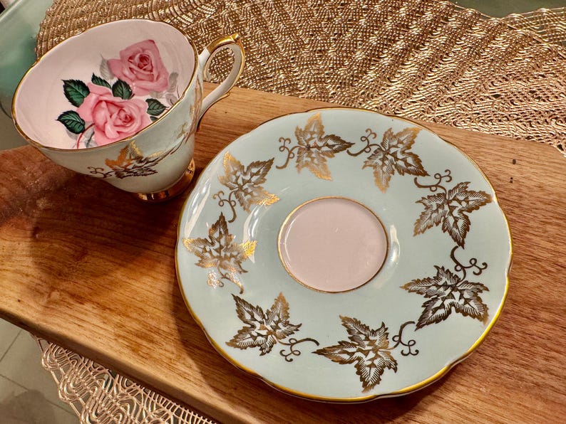 May include: A light blue teacup and saucer set with a gold leaf design. The teacup features a pink rose pattern.