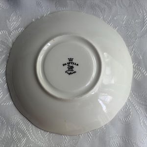 May include: A white ceramic saucer viewed from the underside, featuring a central circular indentation. The center displays a black stamp with a crown icon and the text PASTELLA, ROYAL ALBERT, BONE CHINA, ENGLAND.