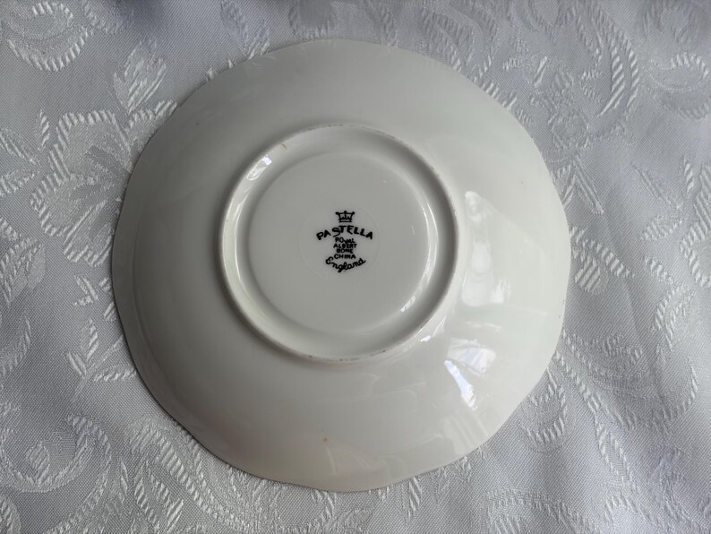 May include: A white ceramic saucer viewed from the underside, featuring a central circular indentation. The center displays a black stamp with a crown icon and the text PASTELLA, ROYAL ALBERT, BONE CHINA, ENGLAND.