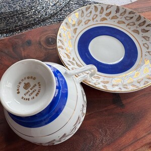 May include: A white teacup and saucer with a blue and gold design. The teacup has a gold rim and the saucer has a gold rim and a gold floral pattern. The teacup has the words "Fine Bone China Made in England" on the bottom.