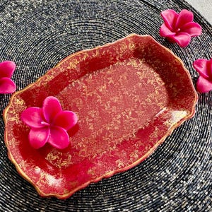 May include: A red and gold decorative tray with a floral pattern. The tray is surrounded by four pink plumeria flowers.