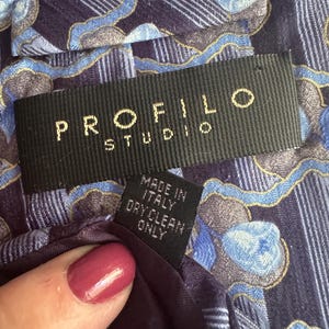 Puede incluir: Una corbata de tela con estampado en azul y morado oscuro con una etiqueta negra que dice PROFILO STUDIO en letras doradas. Una etiqueta negra m&aacute;s peque&ntilde;a adjunta debajo indica MADE IN ITALY y DRY CLEAN ONLY en texto blanco.