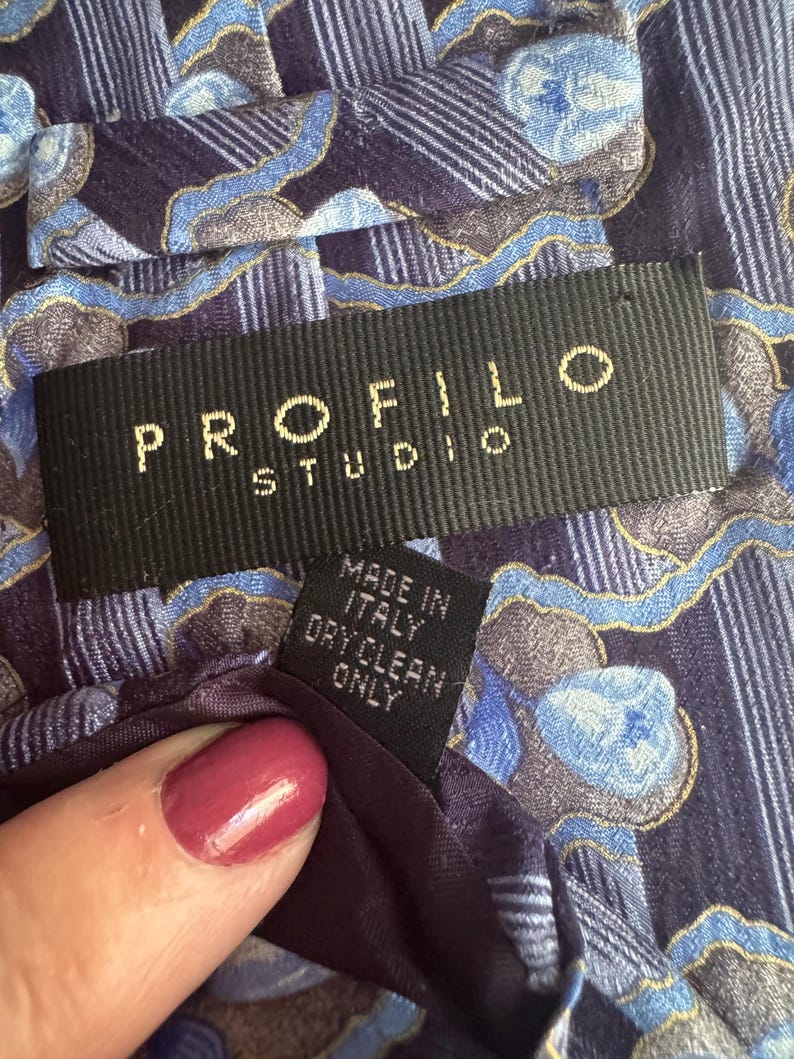 Puede incluir: Una corbata de tela con estampado en azul y morado oscuro con una etiqueta negra que dice PROFILO STUDIO en letras doradas. Una etiqueta negra m&aacute;s peque&ntilde;a adjunta debajo indica MADE IN ITALY y DRY CLEAN ONLY en texto blanco.