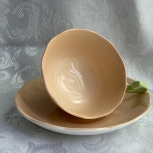 May include: A peach-colored ceramic teacup and saucer set featuring a scalloped edge design. The teacup includes a light green handle, and both pieces have a smooth, glossy finish. The set is presented as a single unit with the cup resting on the saucer.