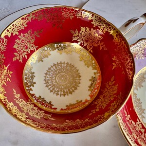 May include: A red and gold floral patterned teacup and saucer set. The teacup and saucer have a scalloped edge and a gold floral design. The saucer is cream-colored with a gold floral design in the center. A gold handle is visible.