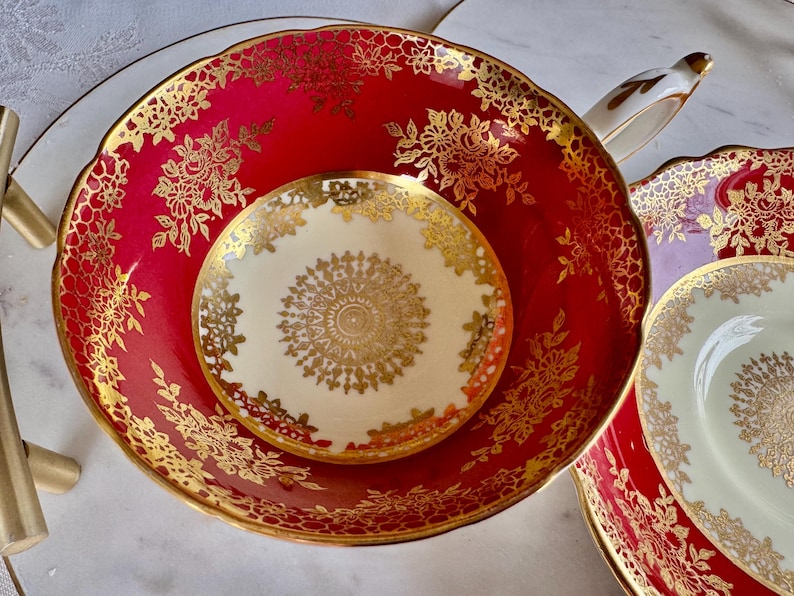 May include: A red and gold floral patterned teacup and saucer set. The teacup and saucer have a scalloped edge and a gold floral design. The saucer is cream-colored with a gold floral design in the center. A gold handle is visible.