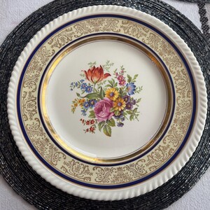 May include: A white porcelain plate with a floral bouquet design. The plate features a gold rim, a blue border, and a cream-colored outer edge with a rope-like texture. The bouquet includes a red tulip, pink rose, and other colorful flowers.