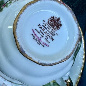 May include: The underside of a white porcelain teacup featuring a gold-trimmed rim and a brown stamp that reads BY APPOINTMENT TO H.M. THE QUEEN CHINA POTTERS PARAGON CHINA LTD. STOKE-ON-TRENT MARLBOROUGH SERIES MADE IN ENGLAND REG'D L/L.