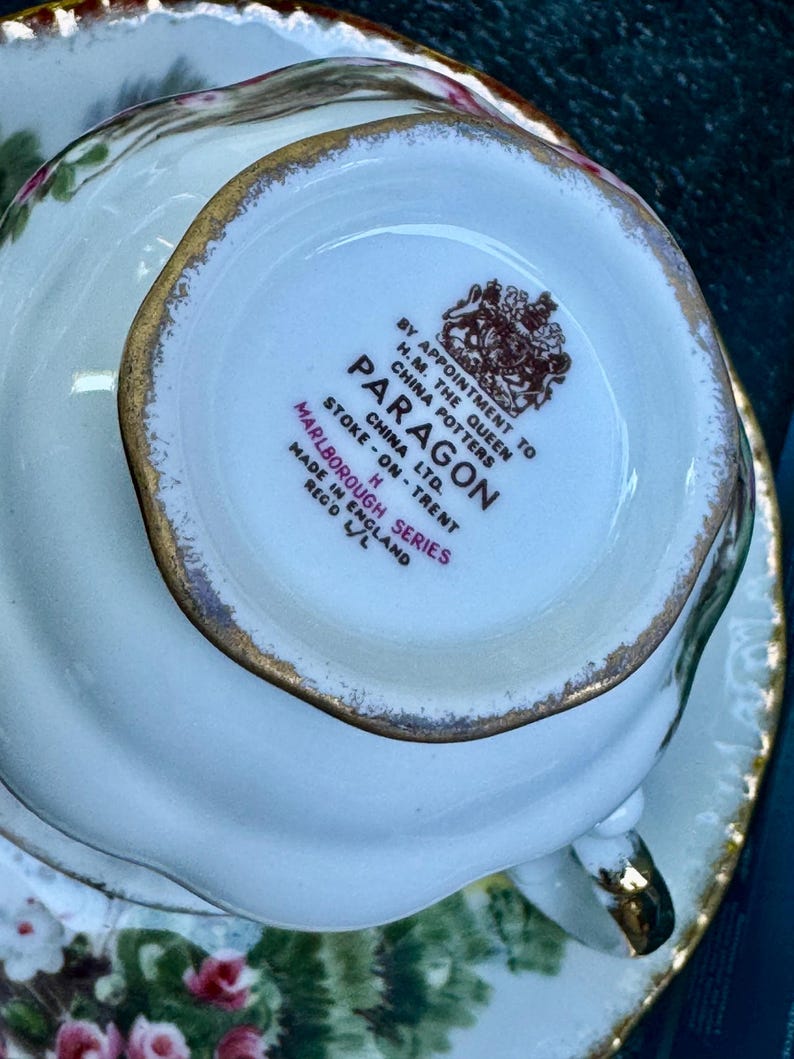 May include: The underside of a white porcelain teacup featuring a gold-trimmed rim and a brown stamp that reads BY APPOINTMENT TO H.M. THE QUEEN CHINA POTTERS PARAGON CHINA LTD. STOKE-ON-TRENT MARLBOROUGH SERIES MADE IN ENGLAND REG'D L/L.