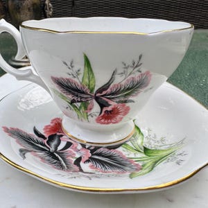 Puede incluir: Juego de taza y platillo blancos con un dise&ntilde;o floral en rosa, negro y verde. La taza y el platillo tienen un borde dorado. La taza tiene un asa curvada y se apoya en un peque&ntilde;o pedestal. El platillo tiene un borde festoneado.