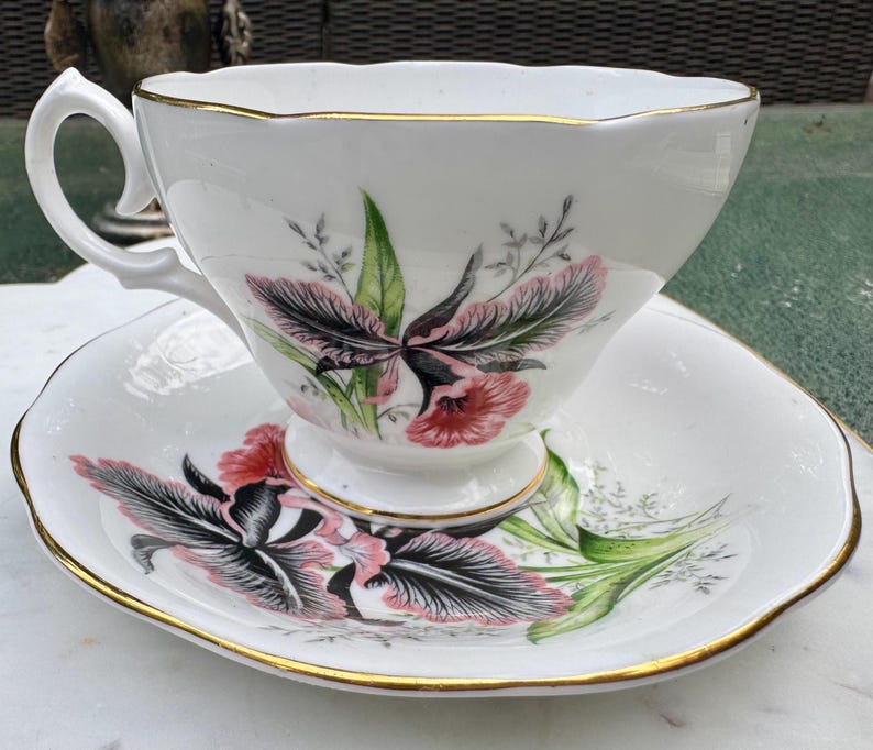 Puede incluir: Juego de taza y platillo blancos con un dise&ntilde;o floral en rosa, negro y verde. La taza y el platillo tienen un borde dorado. La taza tiene un asa curvada y se apoya en un peque&ntilde;o pedestal. El platillo tiene un borde festoneado.