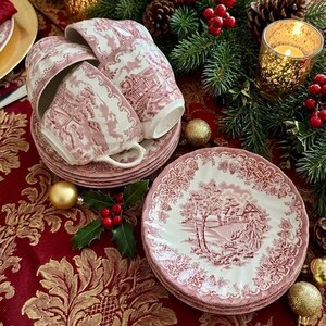 Pu&ograve; includere: Un set di tazze e piattini da t&egrave; in toile bianco e rosso. Le tazze e i piattini presentano un design paesaggistico dettagliato. Il set &egrave; disposto su una tovaglia rossa con accenti dorati, con decorazioni natalizie sullo sfondo.