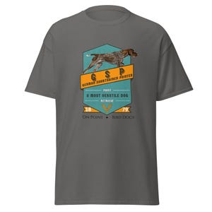 Könnte beinhalten: Ein graues T-Shirt mit einem Aufdruck eines Deutsch Kurzhaar Hundes. Der Aufdruck enthält den Text "GSP", "Deutsch Kurzhaar", "Point", "Ein sehr vielseitiger Hund", "Apportieren", "18", "79", "On Point" und "Bird Dogs".