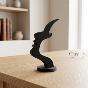 May include: A black abstract sculpture depicting a human profile with a curved, elongated form. The sculpture is on a circular base, placed on a light wooden surface. The background features a bookshelf and a pair of spectacles.