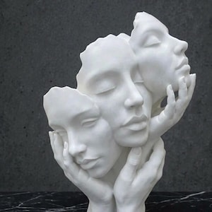 Pode incluir: Uma escultura branca apresentando três rostos humanos interconectados com olhos fechados, sustentados por duas mãos na base. Os rostos de acabamento fosco e liso estão dispostos verticalmente, criando uma forma unificada e abstrata.