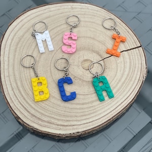 May include: A set of six colourful letter keychains made to look like Lego bricks. The letters are M, S, I, B, C, and A. The keychains are on a wooden surface.