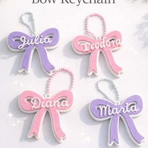May include: Personalised bow keychains in pink and purple with names Julia, Teodora, Diana, and Maria. Each keychain has a white border and a beaded chain. The text "Personalized Bow Keychain" is at the top.