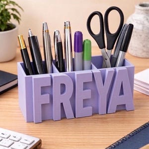 May include: A lilac desk tidy with the name "FREYA" in raised letters. The organiser is divided into three sections, holding pens, markers, and scissors. The desk is made of wood.