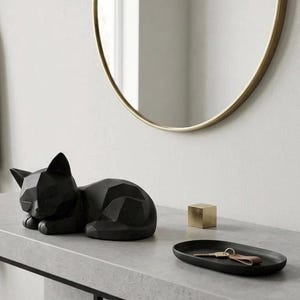 Może przedstawiać: Matowa czarna geometryczna figurka kota w pozycji leżącej, mała złota kostka oraz czarna owalna taca z kluczami. Przedmioty znajdują się na jasnoszarej powierzchni, a nad nimi wisi okrągłe lustro w złotej ramie na ścianie.